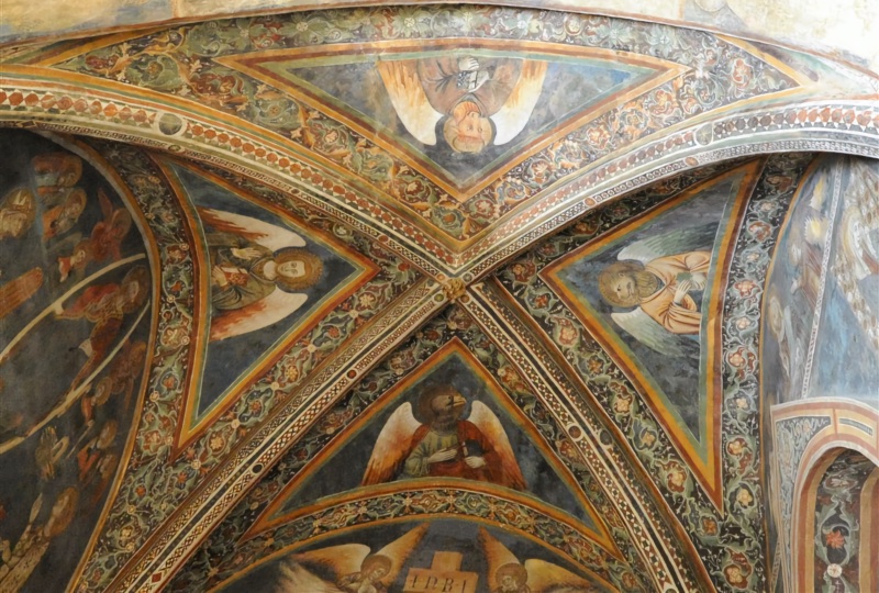 View up into a ribbed cross vault painted in fresco. Four triangular compartments with deep blue grounds each hold a winged, haloed Evangelist with a human body and a book, their heads turned towards the central boss. The figure in the lowest compartment has the head of an eagle, identifying Saint John; the one on the right has a lion's head, the one on the left a human face, and the one at the top, seen upside down, cannot be read with certainty. The ribs carry bands of small geometric motifs in red and white, and broad borders of scrolling foliage and rosettes on a dark ground frame each compartment. At the bottom edge, the top of the Crucifixion with its INRI titulus and two angels is visible; on the left, rows of haloed figures from a side wall; on the right, part of an arched opening