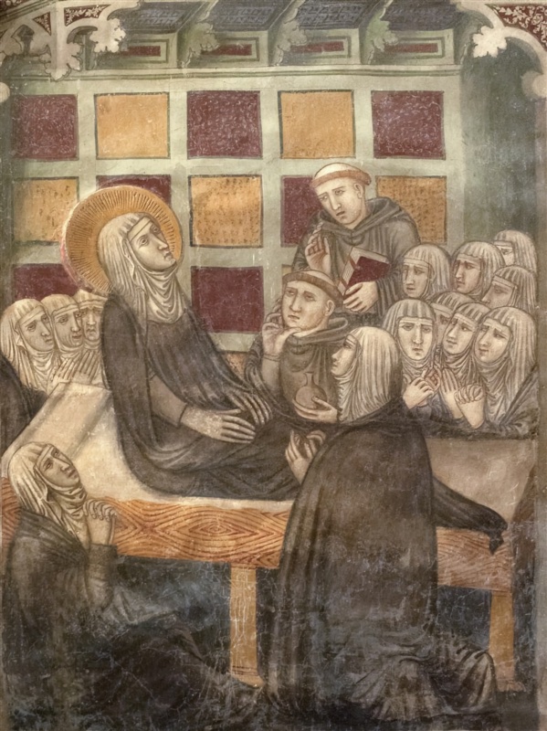 Fresco of a haloed nun in a black habit and white veil sitting upright on a bed, hands folded on her lap, eyes raised to heaven. Behind her a wall of square panels alternates dark red and ochre beneath a painted cornice with Gothic leaf ornament. Crowds of grieving nuns in white veils fill both sides, hands clasped. Two tonsured friars stand beside her: one raises his hand and holds a red book; the other rests his cheek on his hand and holds a small round flask. In the foreground a nun kneels at the bed looking up at her, and another, seen from behind in a dark mantle, kneels at the right. The bed is covered with an ochre cloth patterned in red