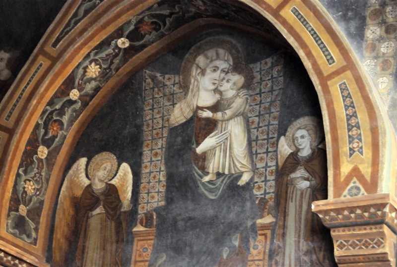 Fresco within a pointed arch showing the Virgin enthroned, wearing a white veil over her head and shoulders and a dark mantle over a red dress. The Christ Child, in a pale tunic and barefoot, stands on her lap, embracing her neck and pressing his cheek to hers. Behind them hangs a cloth of honour with a small red and white chequered pattern. At either side stands a haloed, winged angel in a brown mantle, hands held to the chest. The arch is framed by a band of scrolling foliage with white and orange flowers on a dark ground and an outer border with geometric inlay, resting on a painted corbel with sawtooth ornament