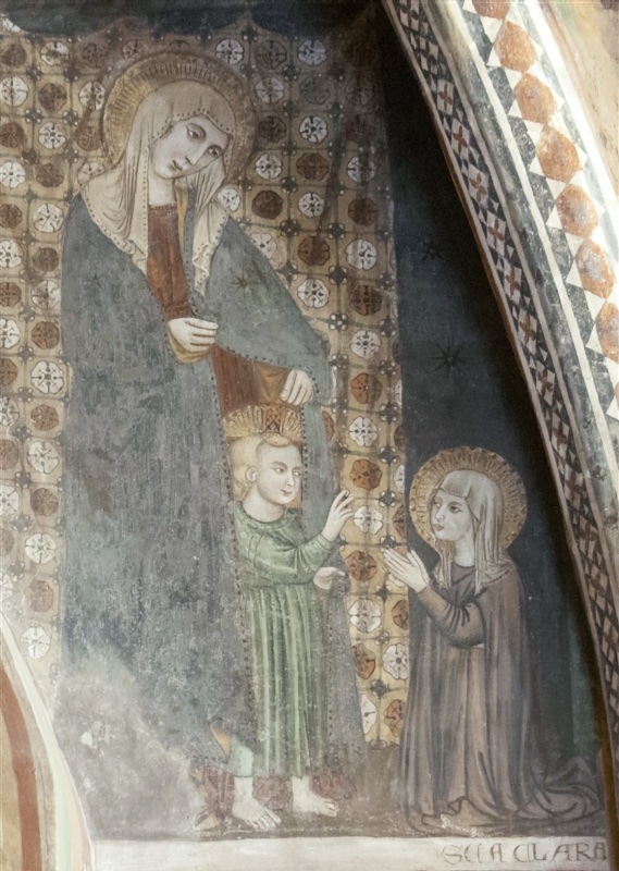 Fresco of a tall standing Virgin in a white veil and grey-green mantle over a red dress, her head bowed towards the right. She draws her mantle open, and from beneath it the barefoot Christ Child, in a green tunic and cruciform halo, steps forward and reaches out his hand. On the right a smaller haloed nun in a brown habit and white veil kneels and raises both hands towards him. Behind the Virgin hangs a cloth patterned with white and red rosettes; to the right is a dark blue ground scattered with stars. An inscription at lower right reads SCA CLARA, and a border of red and white triangles runs along the arch at the edge